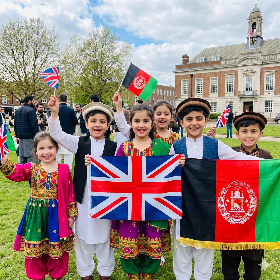 Joyful Afghan children in traditional clothing holding British and Afghan flags on a green lawn in front of a town hall